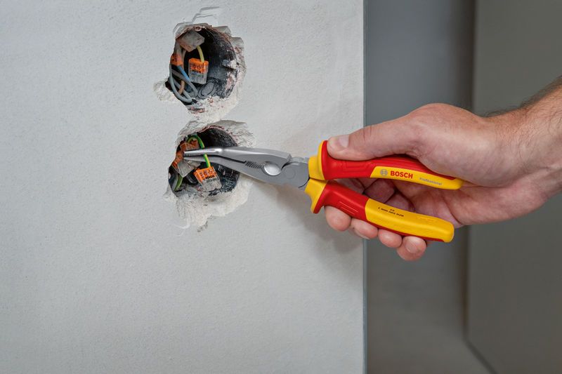 A person uses insulated bent nose pliers to adjust electrical wiring in a wall socket.