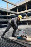 A person wearing safety equipment cuts metal pipes at a construction site with a cut-off saw.