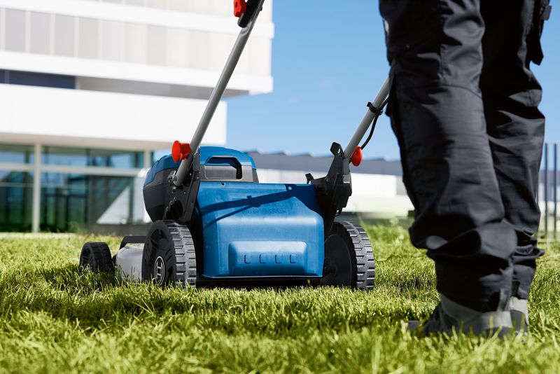 A person wearing safety equipment pushes a cordless lawnmower on green grass.