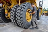 A person wearing safety equipment uses an impact wrench on a large construction vehicle tire.