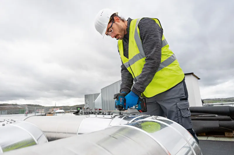 GDR 18V-220 C A person wearing safety equipment uses a power drill on a large metal duct outdoors.