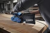 A person wearing safety equipment sands a wooden board with a blue belt sander.