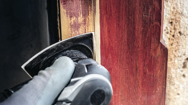 Person wearing safety equipment sands a wooden door frame with a power tool.