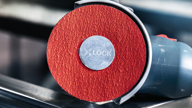 PRO R780 X-Lock Fibre Disc, 125 mm, G36 Close-up of an angle grinder with a red abrasive disc resting on metal.