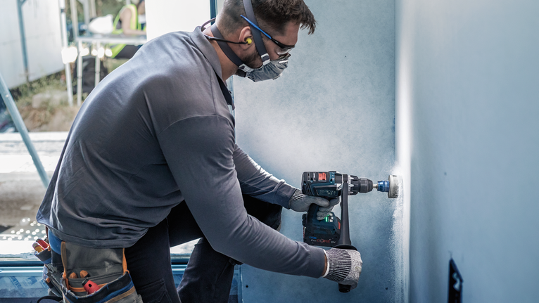 A person wearing safety equipment drills a circular hole in drywall.