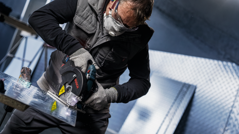 Person wearing safety equipment uses a circular saw to cut a metal sheet.