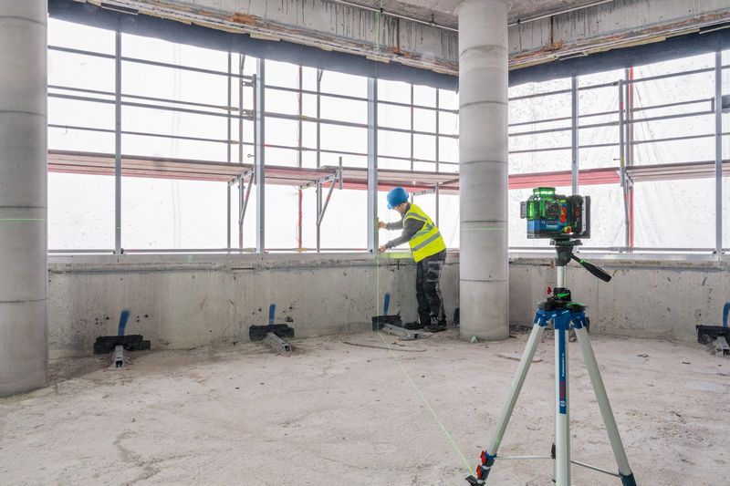 GLL 18V-120-33 CG Person wearing safety equipment aligns materials near a laser leveling tool on a tripod.