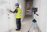 A person wearing safety equipment marks a wall guided by a laser leveling tool on a tripod.