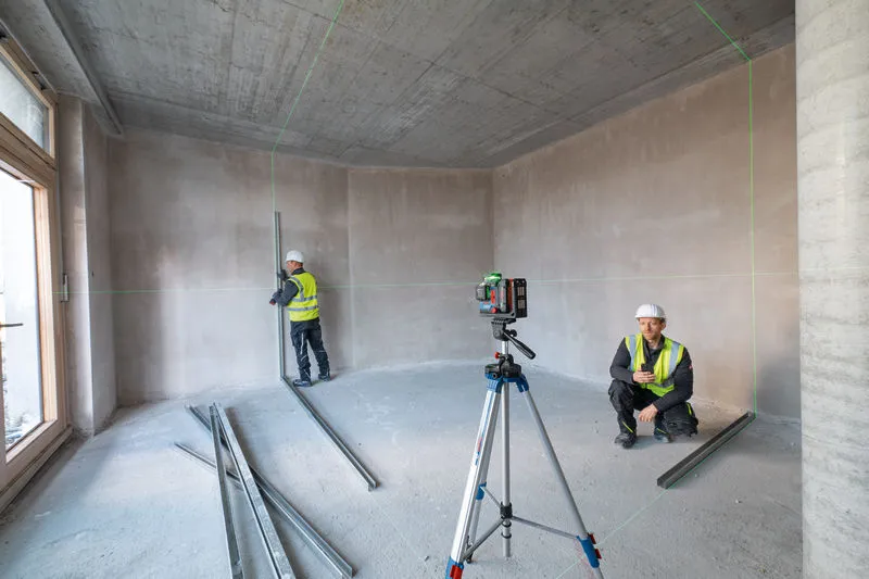 GLL 18V-120-33 CG Two people wearing safety equipment use a laser leveling tool in an unfinished room.