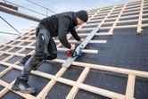 A person wearing safety equipment cuts wood on a steep roof using a cordless circular saw.