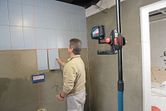 A person aligns wall tiles with a laser leveling tool mounted on a telescopic pole.