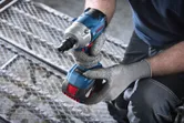 A person wearing safety equipment holds a cordless impact wrench while kneeling on metal grating.