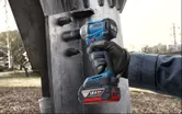 A person wearing safety equipment uses a cordless impact wrench on a large metal structure.