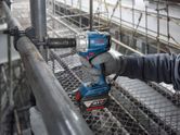A person wearing safety equipment uses a cordless impact wrench on a metal pipe.