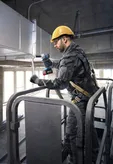 A person wearing safety equipment uses a cordless impact wrench on ductwork.