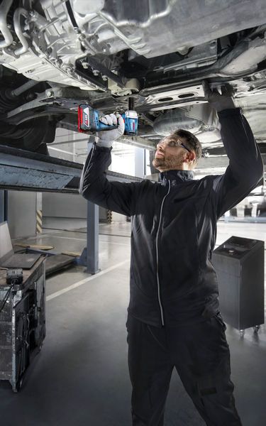 GDS 18V-350 A person wearing safety equipment uses a power drill to work underneath a raised vehicle.