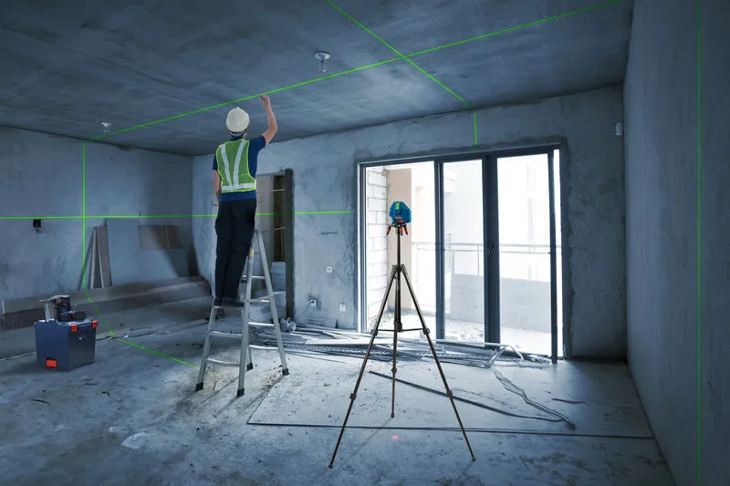 A person wearing safety equipment aligns ceiling fixtures using a laser leveling tool.
