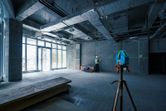 A laser leveling tool on a tripod projects green lines while a person wearing safety equipment marks a wall.