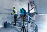 A person wearing safety equipment uses a power drill while a laser leveling tool projects a green line.