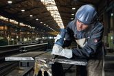 A person wearing safety equipment uses a straight grinder on a metal beam.