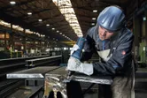 A person wearing safety equipment uses a straight grinder on a metal beam.