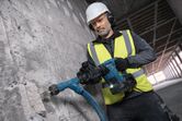 A worker wearing safety equipment uses a cordless demolition hammer on a concrete wall.