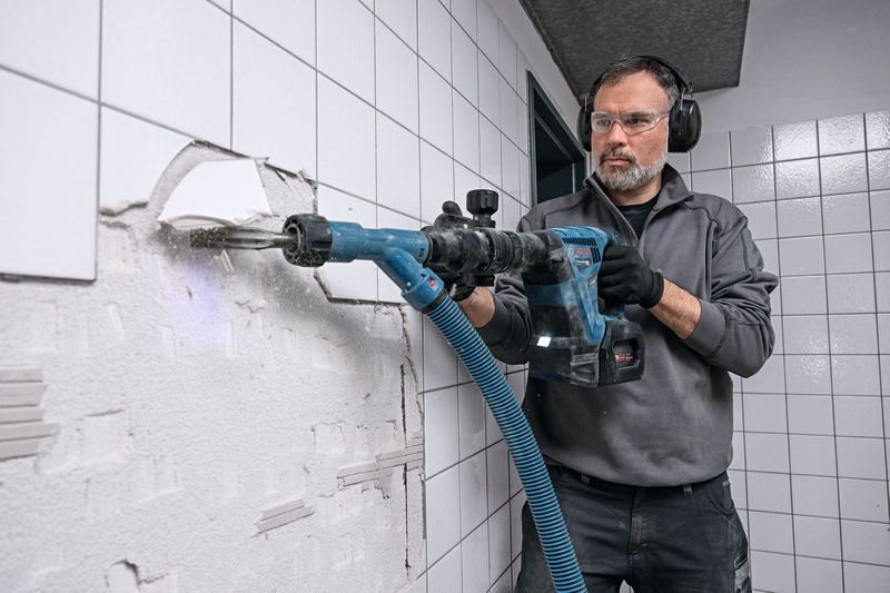 A person wearing safety equipment uses a cordless demolition hammer to break wall tiles.