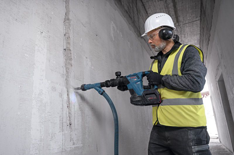 A person wearing safety equipment uses a cordless demolition hammer to break a concrete wall.