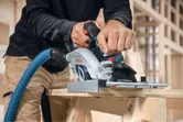 Person cutting wood with a cordless circular saw, connected to a dust extraction hose.