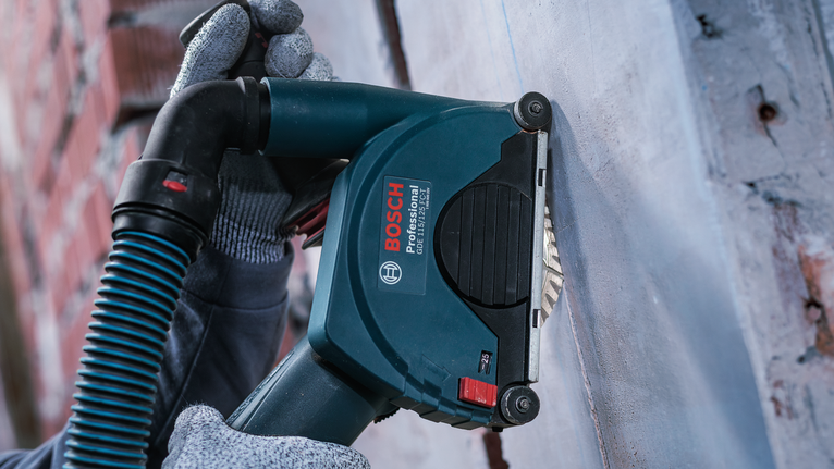 Person wearing safety equipment sands a wall with a power tool.