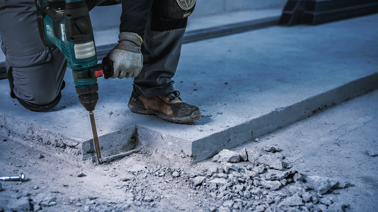 Person wearing safety equipment breaks concrete using a power demolition tool.