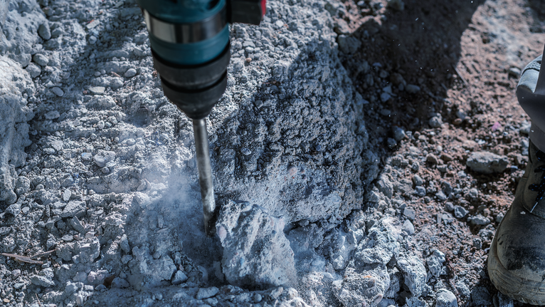 Hammer drill breaking through rough concrete, worker wearing safety equipment.