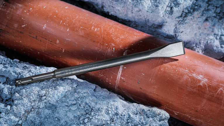 Flat chisel drill bit resting on an orange pipe surrounded by broken concrete.