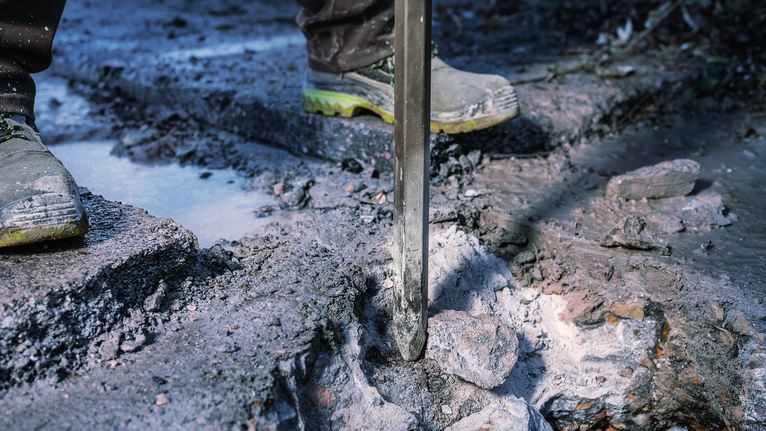 PRO HEX-4C Pointed Chisel 28 mm, 28 x 520 mm A person wearing safety equipment uses a demolition tool to break concrete outdoors.