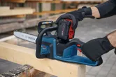 A person wearing safety equipment installs a battery on a cordless chainsaw resting on lumber.