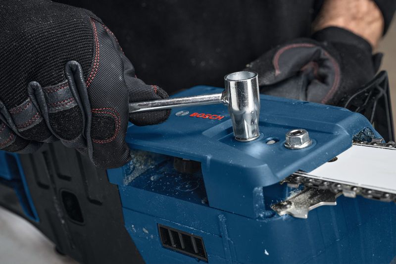 A person wearing safety equipment adjusts a cordless chainsaw with a wrench.
