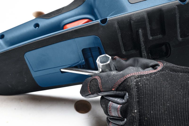 A person wearing safety equipment uses a wrench to adjust a cordless chainsaw.