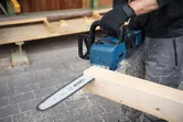 A person wearing safety equipment cuts a wooden beam with a cordless chainsaw.
