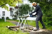 A person wearing safety equipment uses a cordless chainsaw to cut logs outdoors.