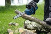 A person wearing safety equipment uses a cordless chainsaw to cut logs outdoors.
