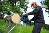 A person wearing safety equipment uses a cordless chainsaw to cut a log outdoors.