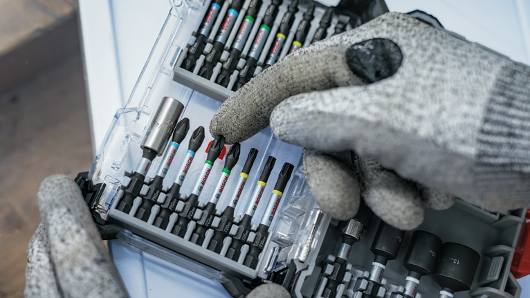 Person wearing safety equipment selects a screwdriver bit from an organized toolkit.