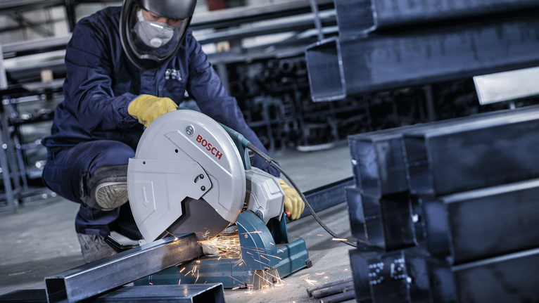 A worker wearing safety equipment cuts metal pipes with a power saw, producing sparks.