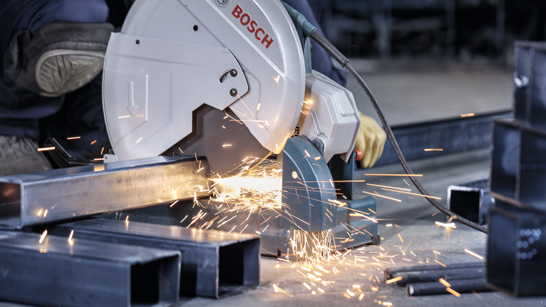 Worker wearing safety equipment cuts metal beams on the floor with a circular saw.