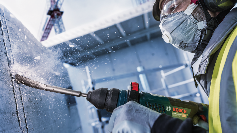 PRO SDS plus Hollow Gouging Chisel, 22 x 250 mm A person wearing safety equipment drills into concrete, creating flying debris.