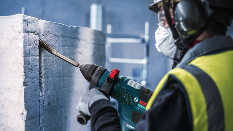 PRO SDS plus Hollow Gouging Chisel, 22 x 250 mm Person wearing safety equipment chisels into a concrete wall with a power tool.