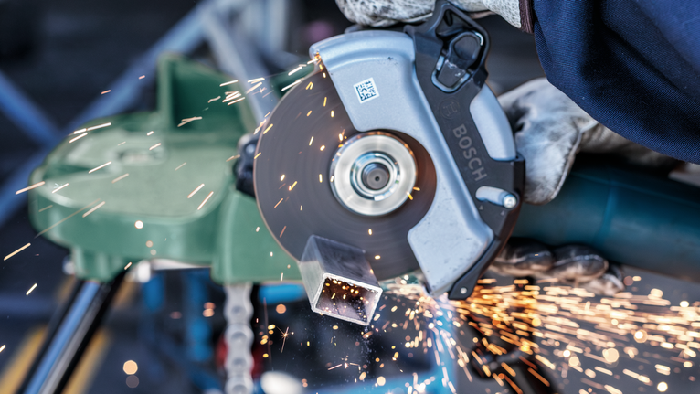 A person wearing safety equipment cuts a metal pipe with an angle grinder, sparks flying.