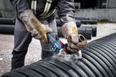 A person wearing safety equipment uses a cordless angle grinder to cut a large plastic pipe.