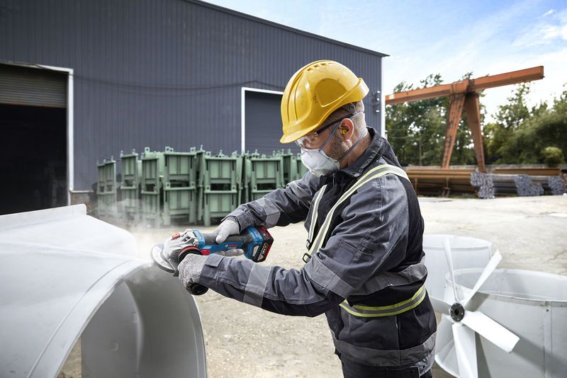 A person wearing safety equipment grinds metal outdoors at an industrial site.