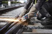 A person wearing safety equipment cuts a railway track with a cordless angle grinder.